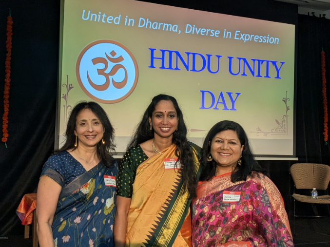 Three women standing in front of a presentation slide that says "United in Dharma, Diverse in Expression. Hindu Unity Day."