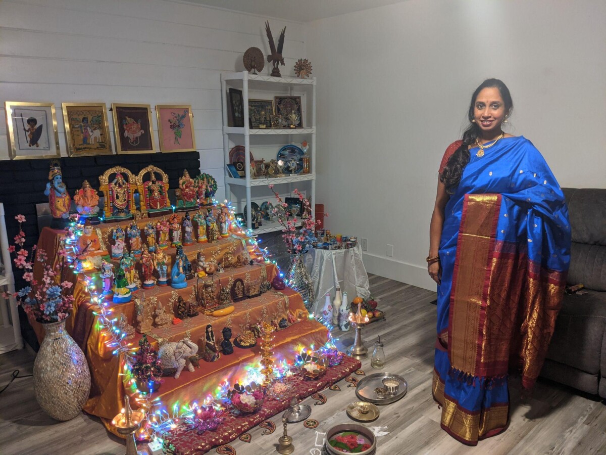 Hindu woman wearing a bright blue silk saree and standing next to a traditional setup of dolls on decorated steps.