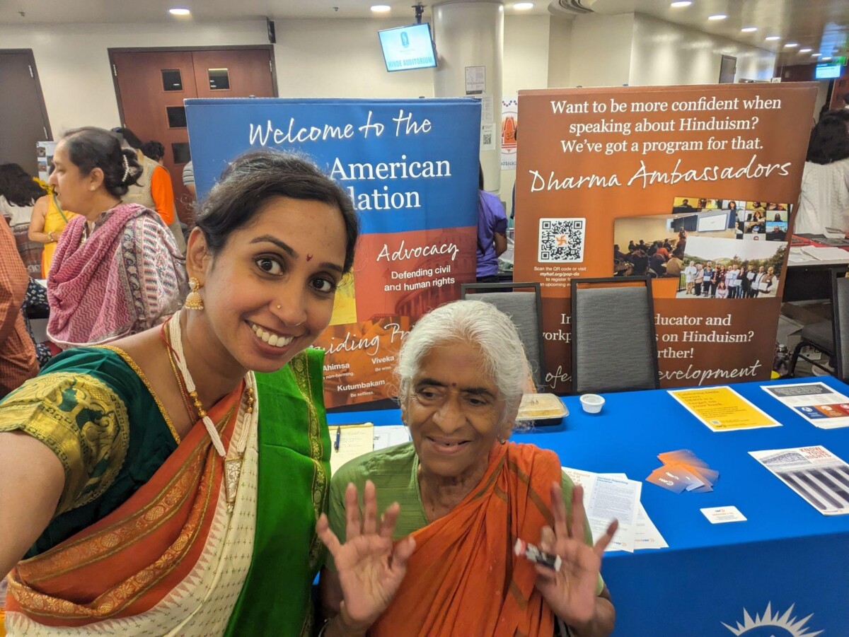 Two Hindu women in sarees pose in front of a HAF booth at a local Sacramento festival